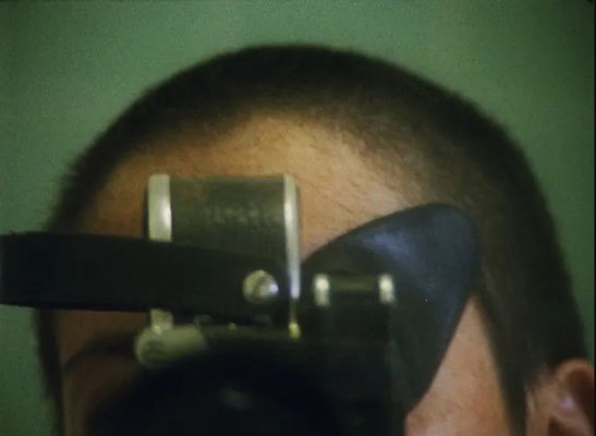 A close-up on someone's forehead and the top of their head. They are holding an object in front of their face. They have light skin and a buzzed head with dark brown hair. The object they are holding resembles a camera and is black and silver.