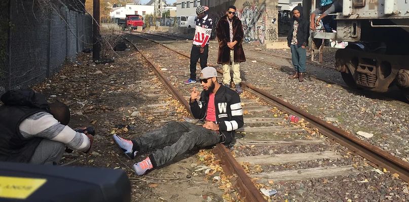 A person with light tan skin sits on the metal edge of a train track in an urban environment. They are wearing a tan baseball cap backwards, a black jersey jacket with a pink t-shirt underneath, black denim pants, and light pink and light blue shoes. There is a person slightly in front of them, hunched over taking a picture of the sitting person with a black camera that is in their hands. The camera-person has dark brown skin, is bald, and is wearing a black puffy sleeveless vest over a white and light-gray-striped long-sleeve t-shirt. In the background, three other people stand around watching the sitting person.