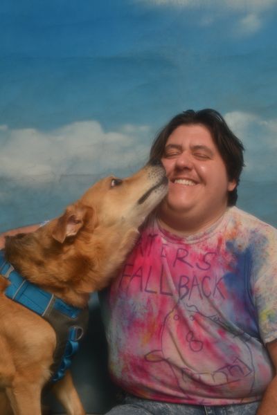 Malik Ever closes their eyes as a lab-sized dog smooches them. Malik is an Algerian artist with light skin, hair falling to their ears, and a tie-dyed t-shirt that likely reads "haters fallback."