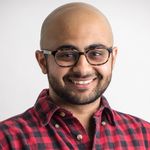 Studio portrait of Anand J. Kalra, a South Asian person. Anand has glasses, brown skin, a short beard and a shaved head. He wears a red and black gingham collared shirt with the top button undone. He is smiling.