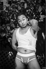 Black and white portrait of Devon Wipp, a Black and Latinx person. Devon has short hair, slight stubble and a couple tattoos. They wear a white tank top pulled up to reveal their stomach and a brief-style strap-on harness with an empty ring. They are outside, standing in front of a chain-link fence penetrated by the leaves of a tree.