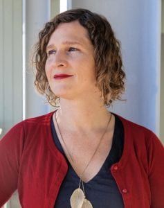 micha cárdenas looks away from the camera with a slight smile. micha is a light-skinned Latinx person, with curly brown-blond chin-length hair. She wears a necklace, v-neck shirt, cardigan and red lipstick.