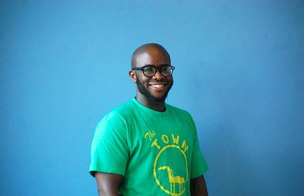 A smiling Shawn Demmons stands in front of a blue background. Shawn wears glasses, is a dark-skinned Black person, and has a shaved head and beard. He wears a green graphic tee that says "The town".