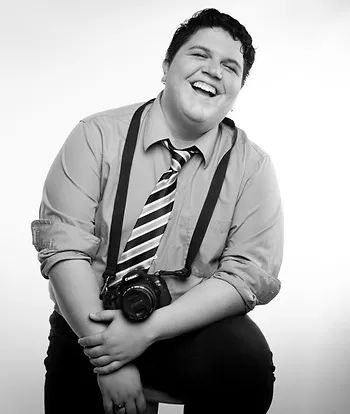Black and white studio portrait of Stephanie Camacho Casillas. Stephanie Camacho Casillas is a Puerto Rican with light skin and shot hair. They wear a button-up shirt, a tie, and have a camera slung around their neck.