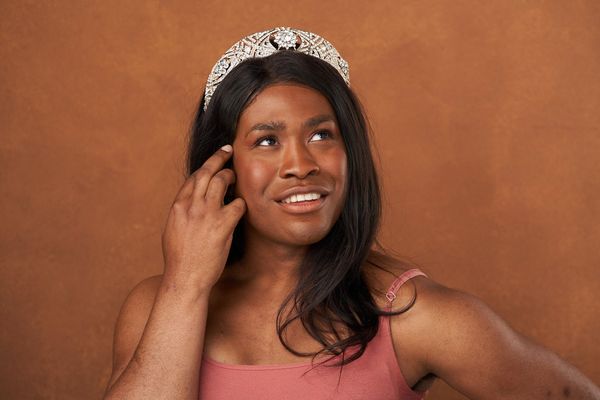 Ava Davis is trans and black with brown skin featured in a shiny stone covered crown and a spaghetti strap tank top. She is looking up and to the right of the image smiling and has her left hand to her face touching her hair. She has dark brown chest length straight hair in front of a brown background.
