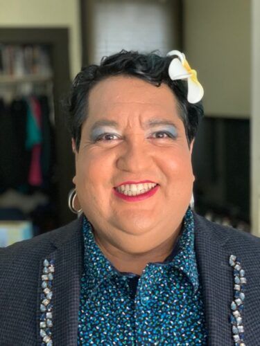 Tina Valentin Aguirre with a big smile. Tina is a Latinx person with short dark hair, light skin and bright makeup. They wear a button-up shirt and blazer.