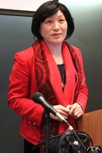 Pauline Park is Korean, wearing a red blazer with a red scarf around their neck and a black shirt underneath. They have light skin and are standing in front of a podium in a classroom with a microphone on the table placed in front of them. They have a black short bob haircut.