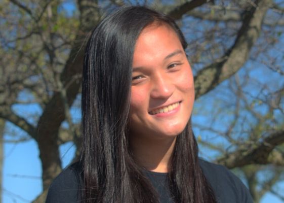 Rey Tang has light skin and is posing outside in front of a tree. She has long straight black hair and a black t-shirt.