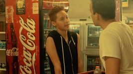 Two people are talking in a convenience store. The person on the left has light tan skin, short light brown hair, and they are wearing a black zip up sleeveless hoodie. They are talking to someone behind the counter. The person behind the counter is wearing a white t-shirt and has light skin and short dark brown hair. Their back is to the camera. The background is a Coca-Cola fridge and some other shelves.
