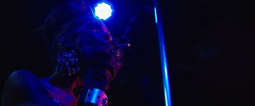 A person with dark brown skin is smoking a cigarette. They have dark brown hair in a big top bun and really big lashes and giant shiny earrings on. They are wearing black velvet gloves with a silver bracelet and a diamond ring on their finger. They have a blue light shining on them and a pole slightly behind them on their right side.