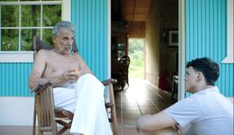 Two people sit out on the porch of a sky-blue house, looking at each other. The person on the right is young, with light tan skin and dark black hair. They are wearing a light gray t-shirt, and are sitting on the ground with their back to the camera, looking up at the person on the left. The person on the left is older, with light tan skin and dark gray hair. They are sitting in a rocking chair, looking down towards the person on the left and speaking to them. They are wearing white pants and no shirt, and are toying with a light orange circular object which could be a ball or some kind of fruit.