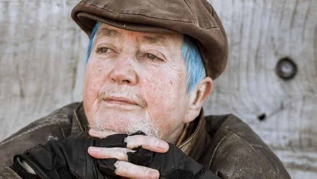 Closeup intimate portrait of Mani slightly turned to the left and gazing at a horizon far away. They are a white intersex person in their 60s with a unique personal style of short light blue hair, natural light brown eyebrows and white facial hair. They are wearing a brown flat cap hat, brown leather jacket and black fingerless gloves. They sit in front of a pale wooden wall.