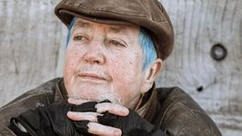 Closeup intimate portrait of Mani slightly turned to the left and gazing at a horizon far away. They are a white intersex person in their 60s with a unique personal style of short light blue hair, natural light brown eyebrows and white facial hair. They are wearing a brown flat cap hat, brown leather jacket and black fingerless gloves. They sit in front of a pale wooden wall.