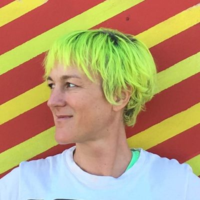 Bright portrait of Lark VCR, a white genderfluid person, against a red-and-yellow diagonal-striped background. They are turned to their right showing their side profile and neon green hair.