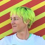 Bright portrait of Lark VCR, a white genderfluid person, against a red-and-yellow diagonal-striped background. They are turned to their right showing their side profile and neon green hair.