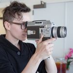 Tommy Statkiewicz holding and looking through the viewfinder of a vintage film camera. He has fair skin, short wavy brown hair, and is wearing glasses and a dark buttoned sweater. The background is a softly-blurred bright and clean room interior.