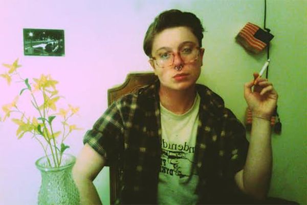 Lauren Soldano in a composed self portrait staring directly into the camera nonchalantly while seated, holding a cigarette in one hand. They are wearing a plaid shirt with rolled sleeves on top of a graphic t-shirt. A vase of yellow flowers is on the table beside them, and a small photo is on the wall in the background. The faded purple-and-green lighting or editing techniques give the image a vintage feel.