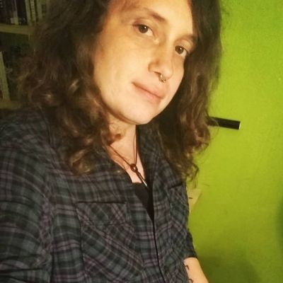 A lo-fi selfie taken by Tylyn S. Anson standing in a room with a bright green wall and bookshelf. She has medium-length curly brown hair, a septum piercing, and wears a plaid shirt.