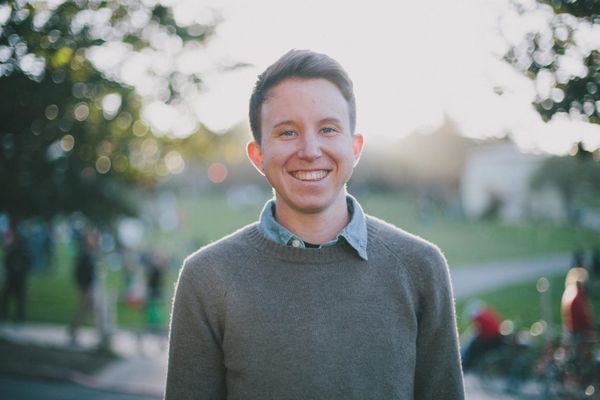 Photograph of Lucas Waldron standing outdoors in a park smiling warmly with a halo-like glow from the sun behind. He is wearing a grew crewneck sweater layered over a blue button up shirt. The background is out of focus and softened, blurring the people along the busy park trails and emphasizing the natural green hues of the trees and grass.