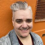 Friendly selfie of Lucas Crawford against a bright orange industrial exterior wall. He has fair skin and short, styled, gray hair. Lucas is wearing a denim jacket and has piercings on his septum, eyebrow and ears.