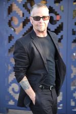 Leon Mostovoy turns his body confidently towards the camera with a slight smile. He is wearing a black shirt under a black blazer. One hand with an arm tattoo is tucked into the pocket of his tailored black pants. He has fair skin, short light hair and facial hair on the chin.