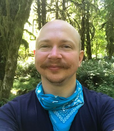 Smiling selfie of Oscar McNary standing in a forest with beams of sunshine filtering through the trees in the background. He has a bald head and a light mustache, and is wearing a bright blue bandana around his neck on top of a navy blue shirt.