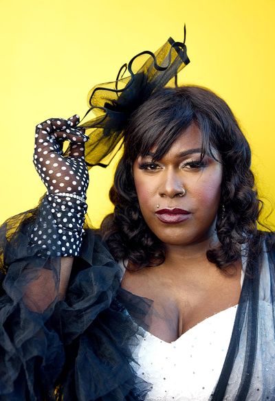 Stylish portrait of Shea Diamond in front of a yellow studio background. Shea is a black trans woman with dark, curly shoulder-length hair. She wears a white dress augmented with dramatic black-lace frills on her arms and head.