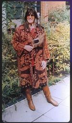 NapéWastéWiñ grins at the camera. She has black hair past her shoulders with bangs. She wears sunglasses, a long umber back with a yellow pattern on it, and brown boots. She holds a large brown bird wing in one hand.