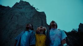 Three adolescents stand at the bottom of a cliff, their arms wrapped around each other, their heads tipped up towards the darkening skies, and their mouths open, perhaps singing or screaming.