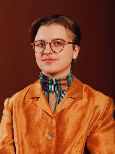 Cassidy Civiero is shown from the chest up in front of a brown studio background. He has round glasses and short brown hair stylishly parted to the sides. Cassidy is wearing a shiny orange suit over a brightly striped multi-color turtleneck.