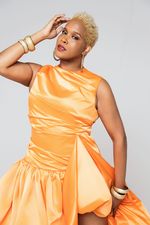 Studio portrait of Neverending Nina with one hand grazing her forehead. She has light brown skin, tight blond curls, and a nose ring. She wears a fancy orange dress and chunky gold bracelets.
