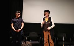 Sam Feder and Kate Bornstein on stage. Sam is a white person with short hair and glasses while Kate is a white person with a blond bob, round sunglasses, and wears a beret. Sam looks at Kate who is holding the microphone and facing the audience.