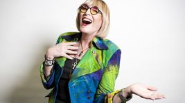 Kate Bornstein with a big laugh and a hand near her chest suggestive of joy. Kate is a white person with a blond bob, round-framed sunglasses, lipstick and nail polish. Kate wears a vibrant jacket of blues and greens over, a punk bracelet and several necklaces.