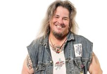 Alec Butler has long sandy hair, a beard, light skin, and a big smile. They are wearing a denim vest with patches, a leather necklace with a pendant, and have some tattoos and piercings.