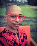 Ignacio G Hutía Xeiti Rivera wears large red glasses, is of Black, Boricua and Taíno descent, and has a shaved head, a couple nose piercings and a slight mustache. They wear a black and red shirt with a geometric print. They are seated on an orange chair with green space behind.