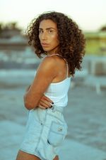 River Gallo stares at the camera over their shoulder with their arms crossed. River is a brown-skinned Salvadorian-American with curly shoulder-length hair. They wear a summer outfit, consisting of a tank top and shorts.