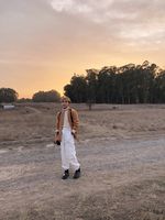 Sal Tran is nestled into a landscape with a road, a field of dried grass, trees, and a sunset sky. Sal Tran is a queer Vietnamese person with edgy short hair. They lazily rest a camera against their thigh and wear a blazer, turtleneck, and cargo pants in a pallet that complements the landscape.