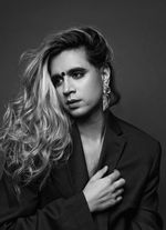 Black and white studio portrait of Vivek Shraya evocative of classic hollywood cinema. She is a South Asian person with tumbling slightly curly hair. She wears a bindi, a chunky earring, and a blazer.