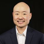 Studio portrait of Alexander L. Lee. Alexander is an East Asian person with a shaved head. He wears a blazer and a collared shirt against a black background. He is smiling.