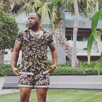 D'Jamel Young stands with his hands resting on his pockets in front of palm trees and a building similar to a hotel. He is a medium-skinned Black person with a beard and short hair. He wears a button-up and matching shorts in the same bold print.