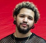 Devin Michael Lowe is a light-skinned Black person with a beard and curly hair. His hair has black roots and blond tips. They wear spiral taper earings, a couple gold chains, and a mesh shirt.