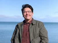 Fallon Simard is an Indigenous person with clear glasses, short black hair, light-skin, and a slight mustache. He wears a light jacket over a button-up shirt. He stands in front of a body of water.