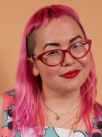 A person smirks into the camera with light skin and bright pink shoulder-length hair with an undercut and bangs. They have red glasses and three stacked gold necklaces. They are wearing a multicolored shirt and red lipstick in front of a peach background.