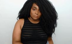 Mahogany Rose looks intently at the camera against a white backdrop. Mahogany is a Black and Indigenous person with medium skin, voluminous dark hair, and a septum peircing. Mahogany wears a fine choker and a black tank top with mesh stripes.