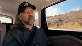 Max Wolf Valerio is inside a car or truck with a serious expression. Max is a Blackfoot and Sephardic person with light skin, short grey hair and beard. He wears a seatbealt, button-up shirt, and baseball cap with a wolf on it.