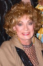 Holly Woodlawn, an older light-skinned Puerto Rican woman with curled golden blonde hair, wears red lipstick, long gold earrings, and a sweater.