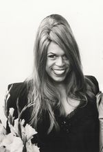 Black and white photo of Vaginal Davis with a big smile. She is a Black and Hispanic person with long straight hair. She stands behind some gladiolus and wears a blazer over a top with lace edging.