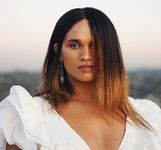 Davia Spain is a Black Trans Woman with light brown skin. She is shown from the chest up in a white v neck top. Davia has dark brown ombre hair covering half of her face. She is wearing long dangly earrings. She is in front of a blurred out landscape during the day time.
