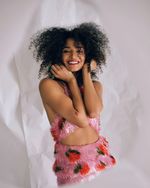 Indya Moore standing with her chin resting on her hands and a smile. Indya is a light-skinned Black person with big curls and a star tattoo near her elbow. She wears a pink halter dress made of fringe with a fruit print.