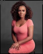 Studio portrait of Janet Mock sitting with a slight smile. Janet is a light-skinned Black person with curly shoulder length hair. She wears a pink form-fitting dress, hoop earings, and a ring on her wedding finger.